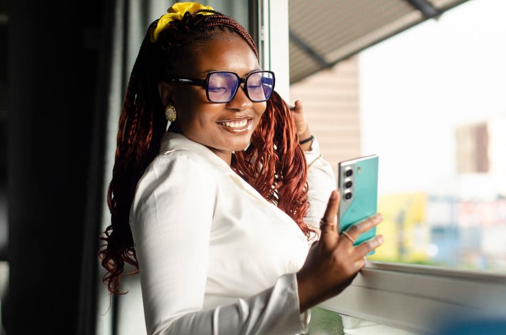 A woman smiling at her phone by a window